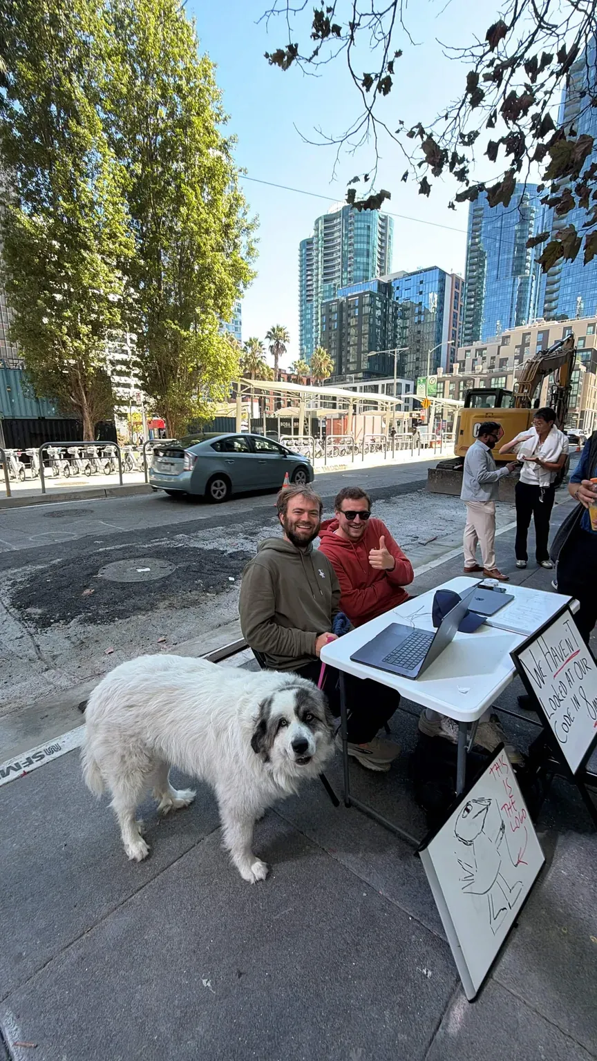 A big fluffy white dog walks past the stand; two of the founders sit behind it in the olive and rust hoodies.